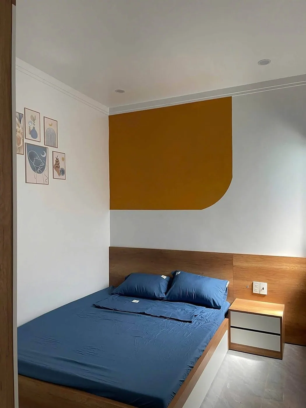 Phòng ngủ đầu giường gỗ, chăn ga xanh, mảng tường cam và tranh trang trí
