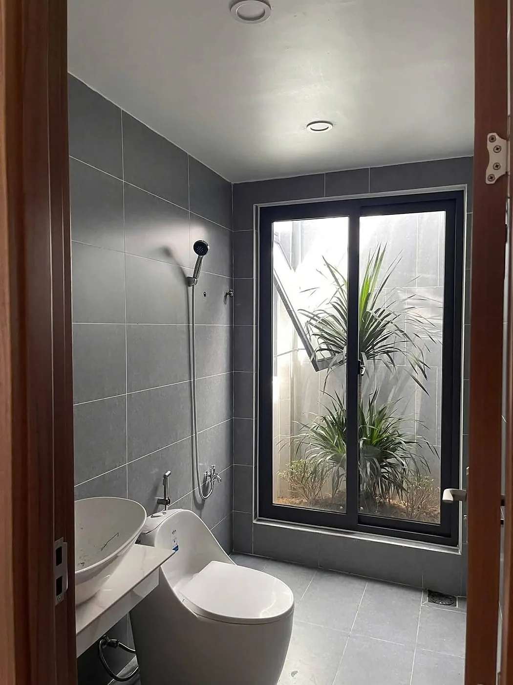 Nhà vệ sinh lát đá xám, cửa kính lớn nhìn ra giếng trời cây xanh