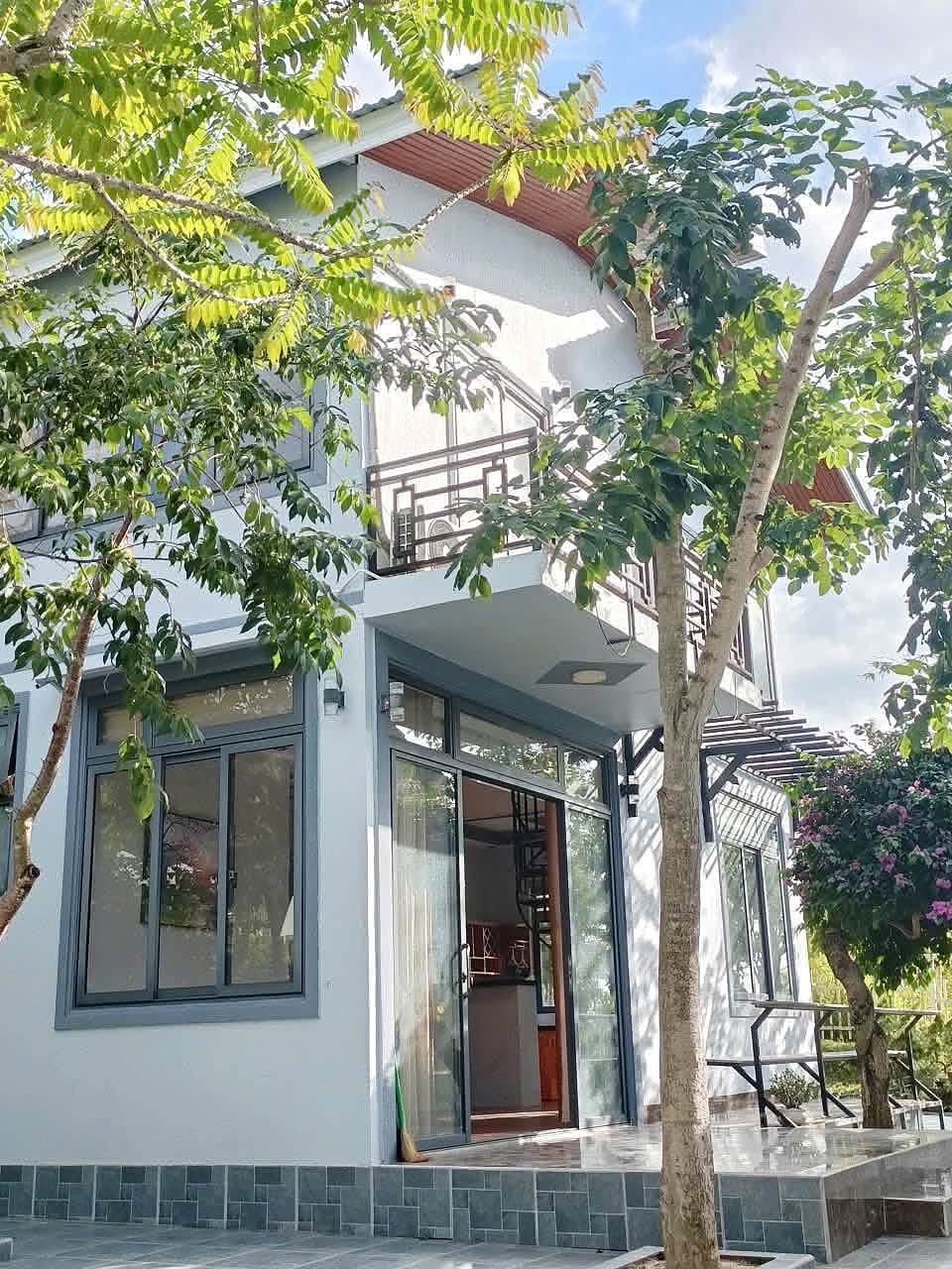 Mặt tiền nhà 2 tầng ở Gia Lâm, Nam Ban: tường trắng, cửa kính khung xám, ban công lan can gỗ, cây xanh trước nhà
