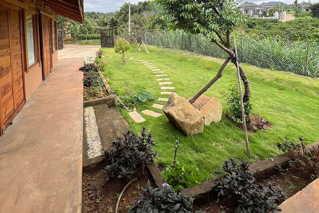 Lối đá dạo qua sân cỏ nhìn từ hiên nhà gỗ, cây xanh và tiểu cảnh đá