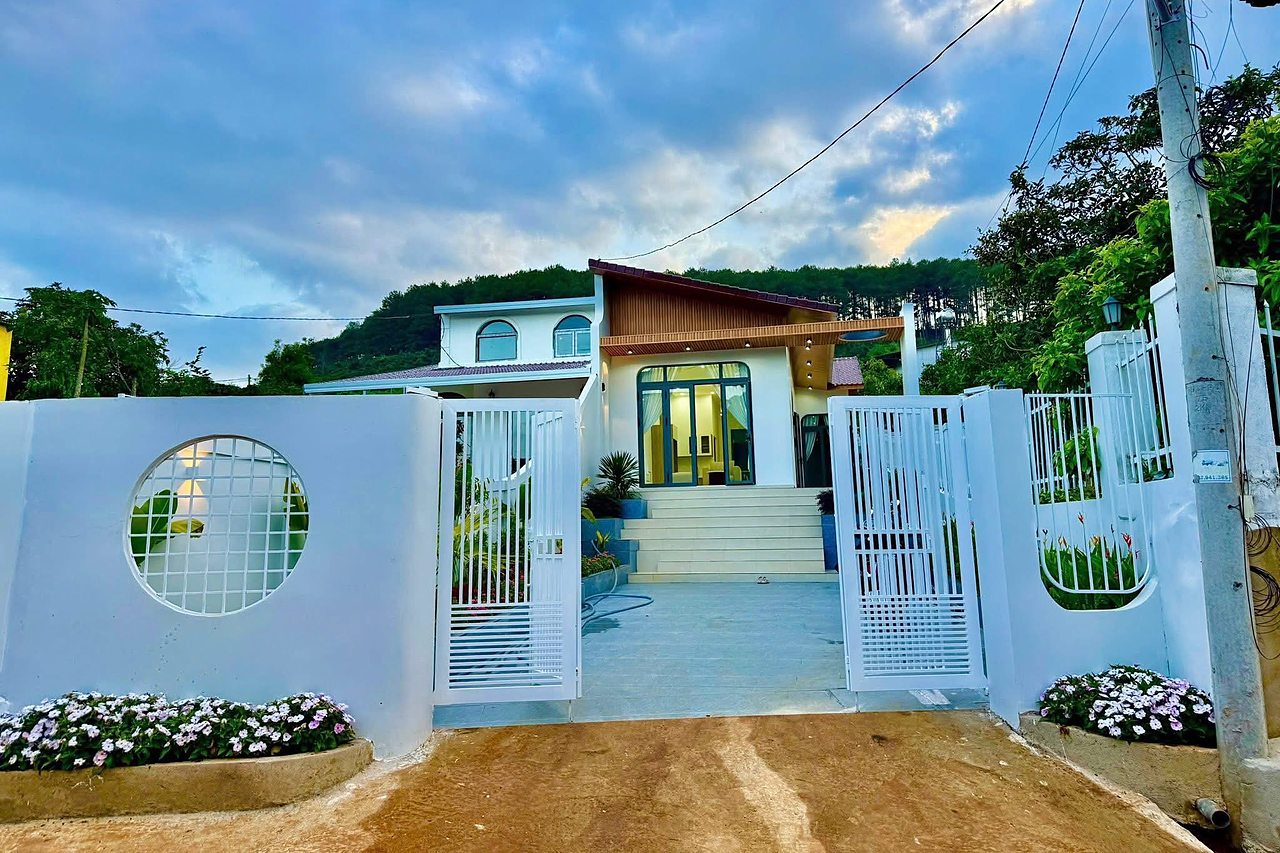 Nhà mới Mê Linh nhìn từ cổng — nhà mái lệch, cửa kính lớn lên đèn, bậc thang sân, tường rào trắng, đồi thông phía sau