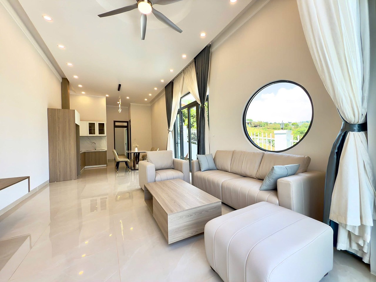 Phòng khách nhà Mê Linh — sofa da kem, cửa sổ tròn view đồi, quạt trần, thông sang bếp, sàn gạch bóng