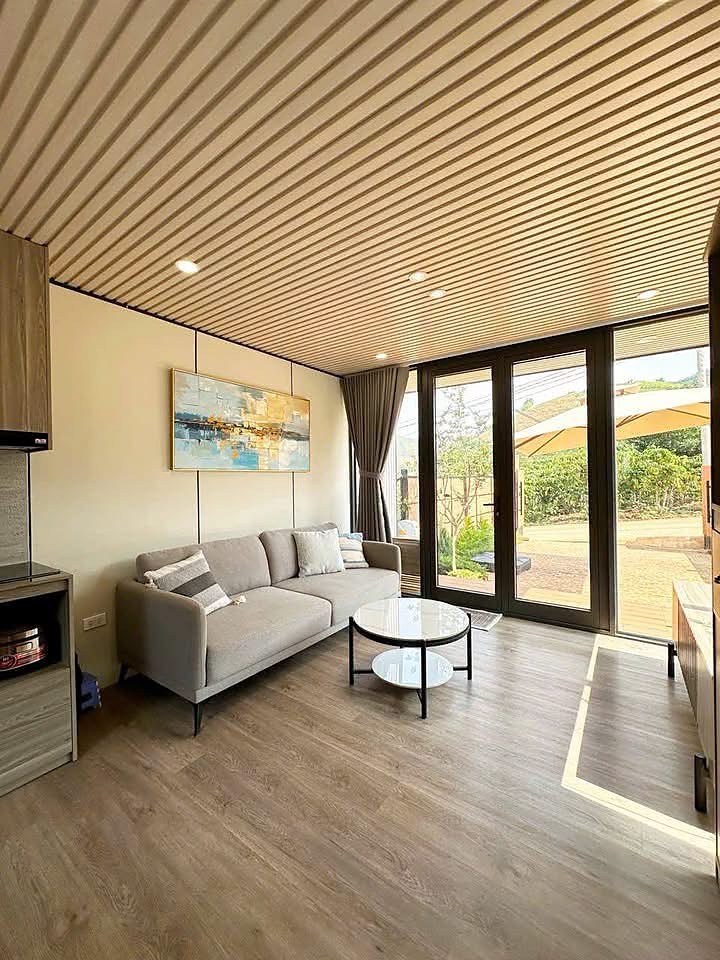 Phòng khách nhà nghỉ dưỡng Mê Linh 189m²: sofa, bàn trà, trần lam gỗ, cửa kính lớn mở ra sân