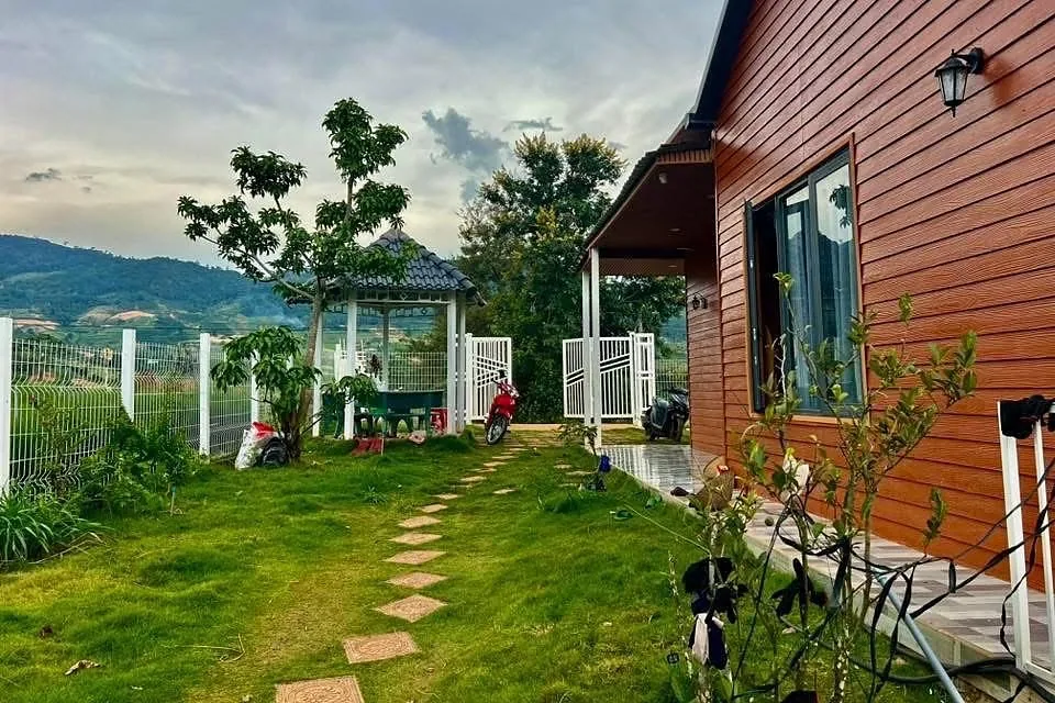 Sân vườn nhà nhỏ với chòi nghỉ và lối đá dạo, tường gỗ đỏ, view đồi lúc chiều