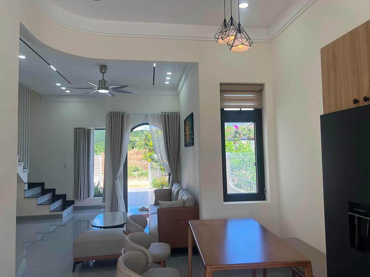Phòng khách liền bàn ăn: sofa, quạt trần, đèn thả, cửa vòm mở ra sân trước, cầu thang lên lầu