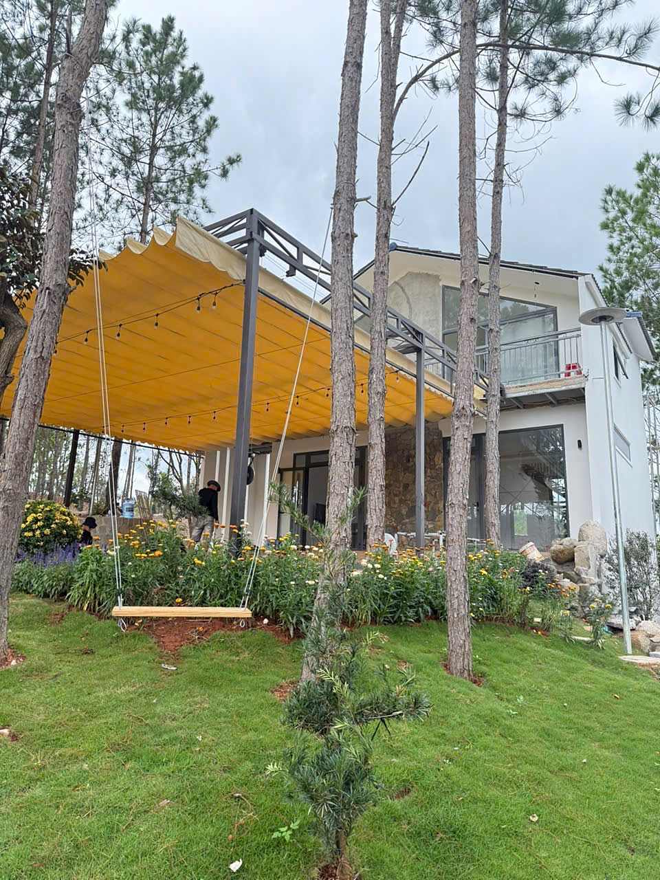 Villa HT86 Nam Ban với mái hiên bạt, đèn dây và vườn hoa, xích đu gỗ dưới tán thông