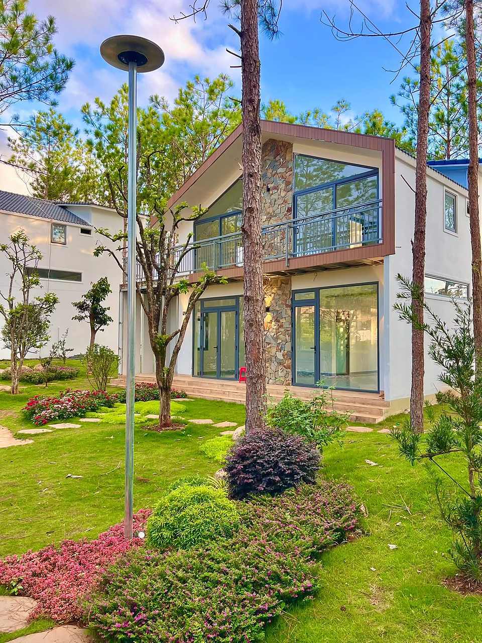 Villa HT86 Nam Ban nhìn từ vườn – cột đá ốp, ban công kính, đèn sân vườn dưới tán thông