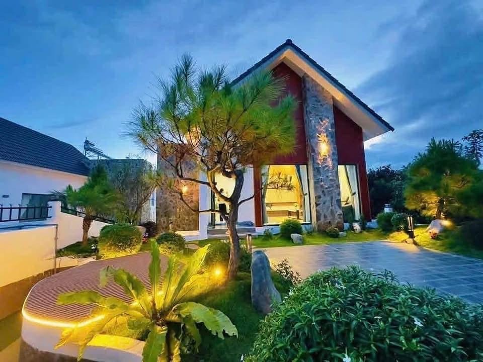 Villa nghỉ dưỡng Nam Ban lên đèn buổi tối – mái dốc, mặt đá và gỗ đỏ, sân vườn cây thông, đèn vàng ấm